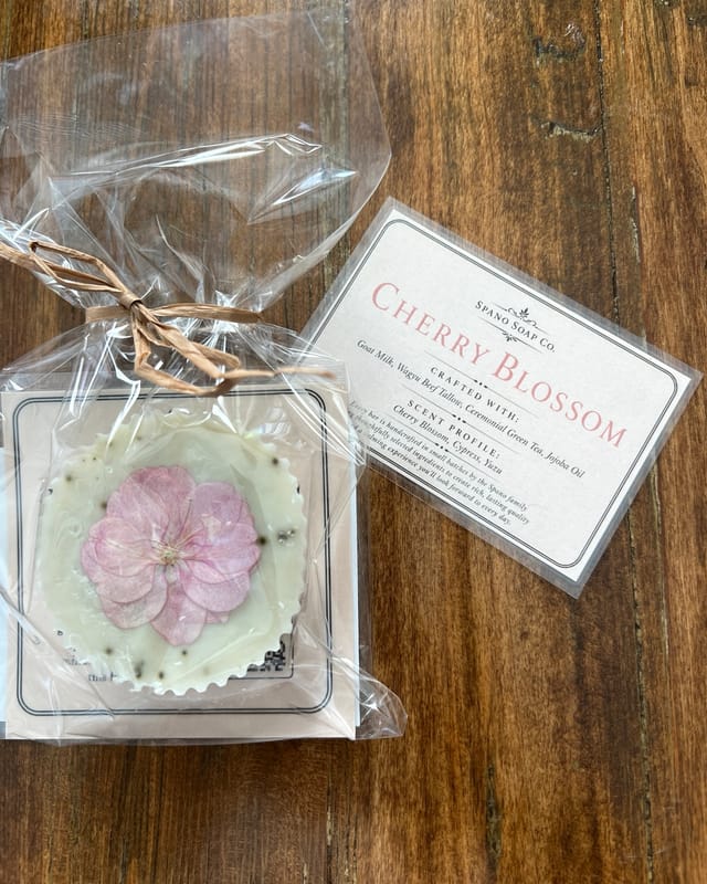 A square Cherry Blossom bar with a pressed pink blossom set in its surface, wrapped in cellophane and tied with raffia, resting on a dark walnut table beside its printed label.