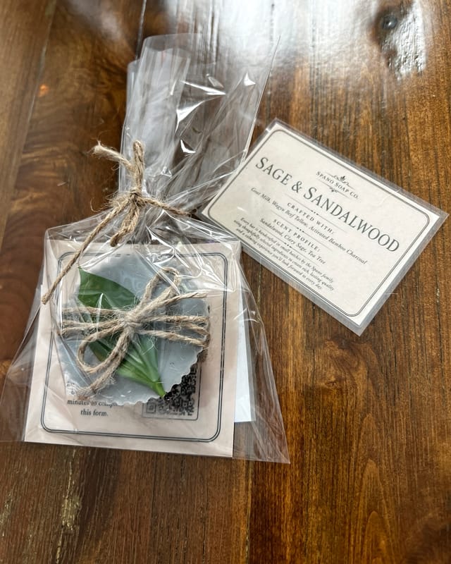 A grey Sage and Sandalwood bar with a green sage leaf laid across it, wrapped in cellophane and tied with garden twine, on a dark walnut table beside its printed label.