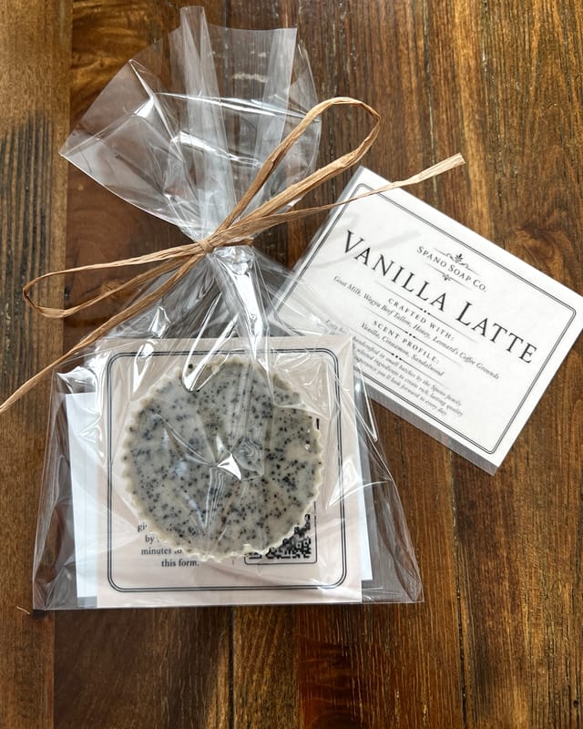 A pale Vanilla Latte bar speckled with dark coffee grounds, wrapped in cellophane and tied with raffia, on a dark walnut table beside its printed label.